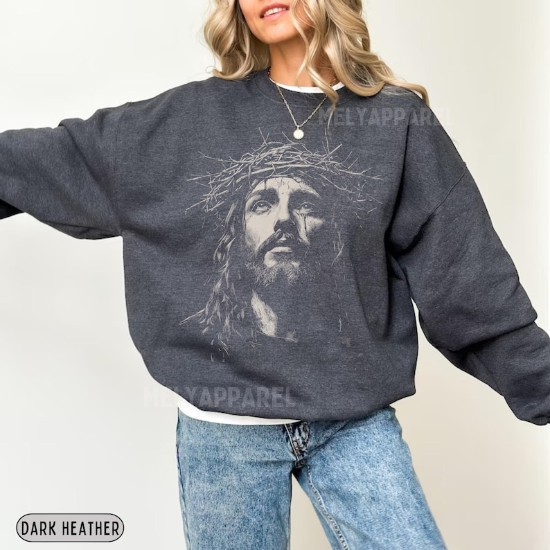May include: Dark heather gray sweatshirt featuring a detailed graphic of Jesus wearing a crown of thorns. The image is centered on the front of the sweatshirt. The text "MELY APPAREL" is printed on the sweatshirt. The words "DARK HEATHER" are printed on the bottom left.