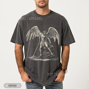 May include: Dark grey t-shirt with a black and white illustration of an angel with large wings and a sword. The text "MELY APPAREL" is printed on the shirt. The shirt is labeled "PEPPER".