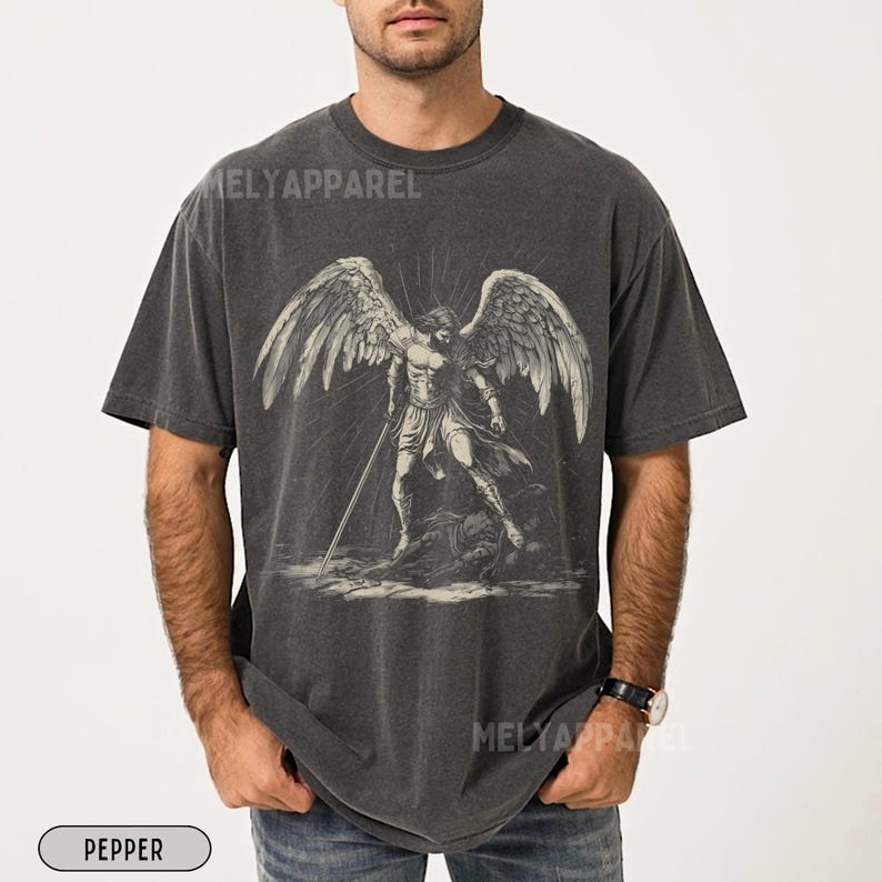 May include: Dark grey t-shirt with a black and white illustration of an angel with large wings and a sword. The text "MELY APPAREL" is printed on the shirt. The shirt is labeled "PEPPER".