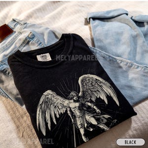 May include: Black crew neck t-shirt featuring an angel graphic with large wings and a sword. The shirt has the text "MELY APPAREL" printed on it. The shirt is folded on top of a pair of light blue jeans.