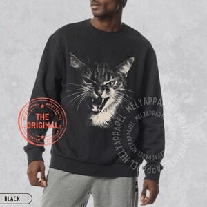May include: A black long-sleeve sweatshirt featuring a white graphic illustration of a hissing cat with bared teeth on the chest. The garment includes a circular red stamp with the text THE ORIGINAL and the repeated text MELYAPPAREL.