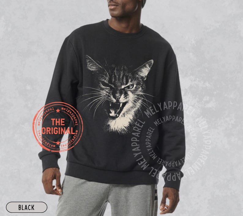 May include: A black long-sleeve sweatshirt featuring a white graphic illustration of a hissing cat with bared teeth on the chest. The garment includes a circular red stamp with the text THE ORIGINAL and the repeated text MELYAPPAREL.