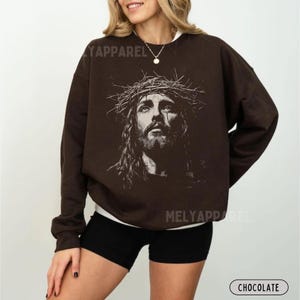 May include: Chocolate-colored sweatshirt featuring a detailed black and white portrait of Jesus wearing a crown of thorns. The sweatshirt has the text "MELY APPAREL" printed on it. The word "CHOCOLATE" is printed on the bottom right.