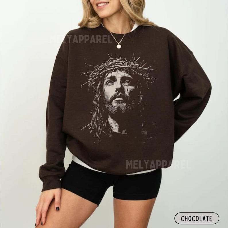 May include: Chocolate-colored sweatshirt featuring a detailed black and white portrait of Jesus wearing a crown of thorns. The sweatshirt has the text "MELY APPAREL" printed on it. The word "CHOCOLATE" is printed on the bottom right.