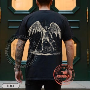 May include: Black t-shirt featuring a detailed illustration of an angel with large wings, holding a sword. The graphic is in shades of gray and white. The shirt has a round neck and short sleeves. The text "THE ORIGINAL" is printed on the bottom right.