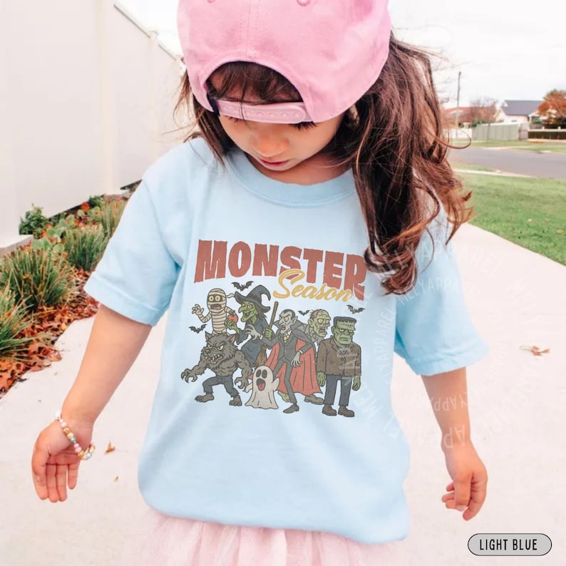 May include: A light blue t-shirt featuring a graphic print of various classic monsters, including a mummy, witch, vampire, werewolf, ghost, and Frankenstein's monster, with the text "MONSTER Season" printed in brown and yellow above the characters.