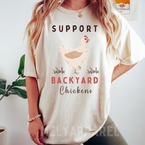 以下が含まれることがあります： 「SUPPORT BACKYARD CHICKENS」という文字と、鶏の漫画の絵が描かれた白いTシャツです。