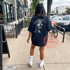 May include: Black oversized t-shirt featuring a detailed grayscale portrait of Jesus wearing a crown of thorns. The shirt has the words "AREUMEL" printed vertically on the left side. A red stamp with the words "THE ORIGINAL" is visible.