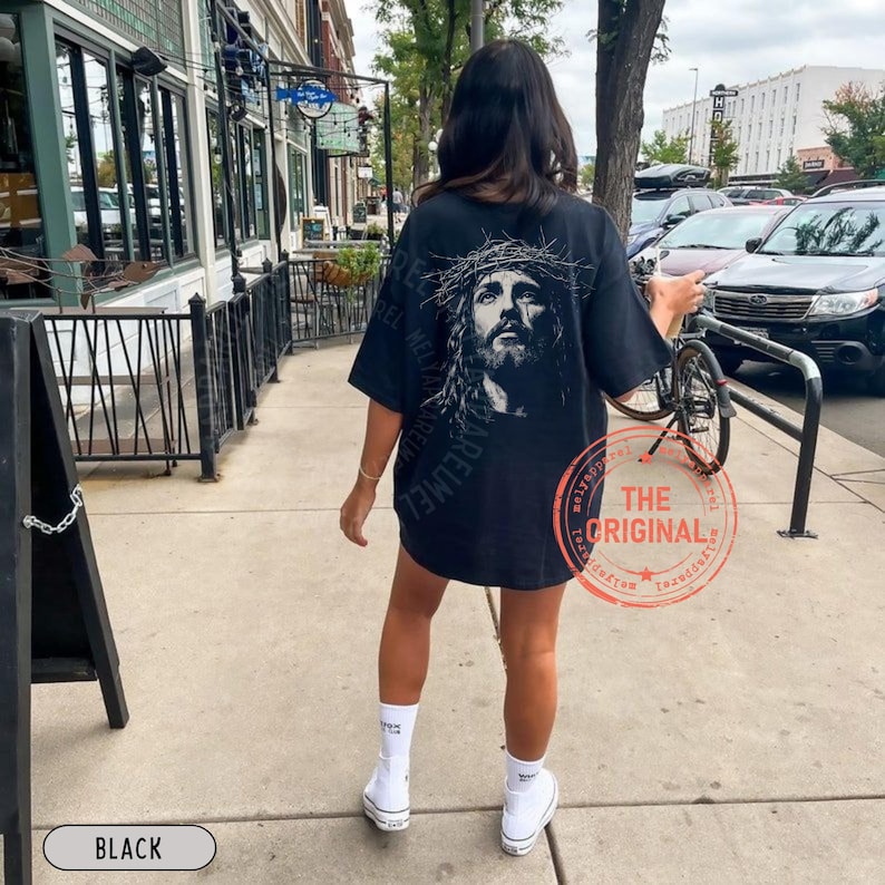 May include: Black oversized t-shirt featuring a detailed grayscale portrait of Jesus wearing a crown of thorns. The shirt has the words "AREUMEL" printed vertically on the left side. A red stamp with the words "THE ORIGINAL" is visible.