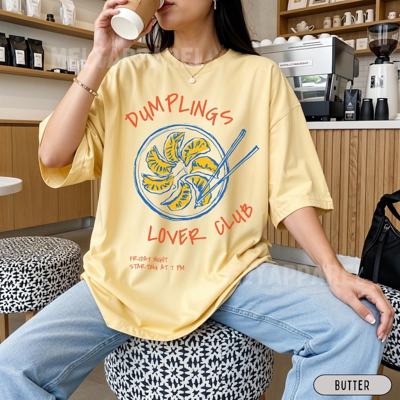 Op de afbeelding: Lichtgeel T-shirt met de tekst "DUMPLINGS LOVER CLUB" in rood en wit. Het shirt heeft een afbeelding van dumplings met eetstokjes. De tekst "FRIDAY NIGHT STARTING AT 7 PM" is ook zichtbaar.