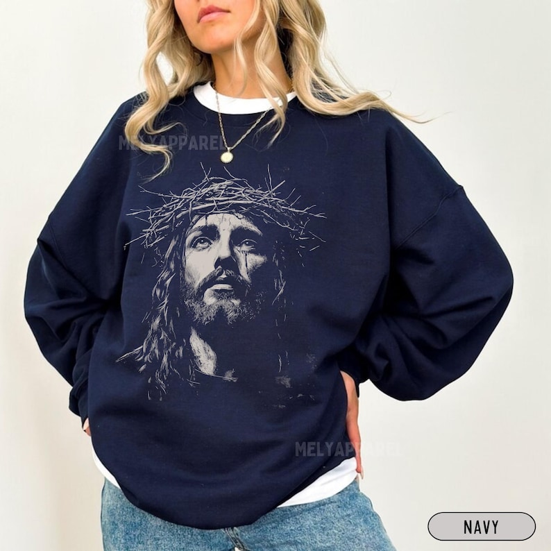 May include: Navy blue sweatshirt featuring a black and white graphic of Jesus wearing a crown of thorns. The sweatshirt has long sleeves and a crew neck. The text "MELY APPAREL" is printed on the upper left and lower right of the sweatshirt. The word "NAVY" is printed on the lower right.