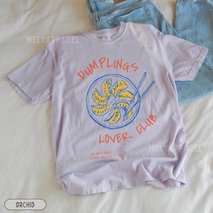 Op de afbeelding: Orchidee-kleurig T-shirt met een afbeelding van dumplings in een blauwe kom met eetstokjes. De tekst luidt "DUMPLINGS LOVER CLUB" in oranje en blauw. Daaronder staat "FRIDAY NIGHT STARTING AT 7 PM".