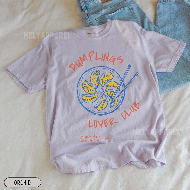 Op de afbeelding: Orchidee-kleurig T-shirt met een afbeelding van dumplings in een blauwe kom met eetstokjes. De tekst luidt "DUMPLINGS LOVER CLUB" in oranje en blauw. Daaronder staat "FRIDAY NIGHT STARTING AT 7 PM".