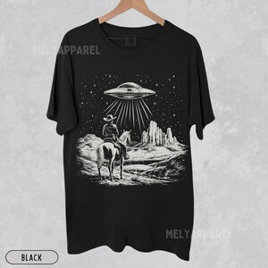 May include: Black t-shirt with a graphic of a cowboy riding a horse under a UFO in a desert landscape.