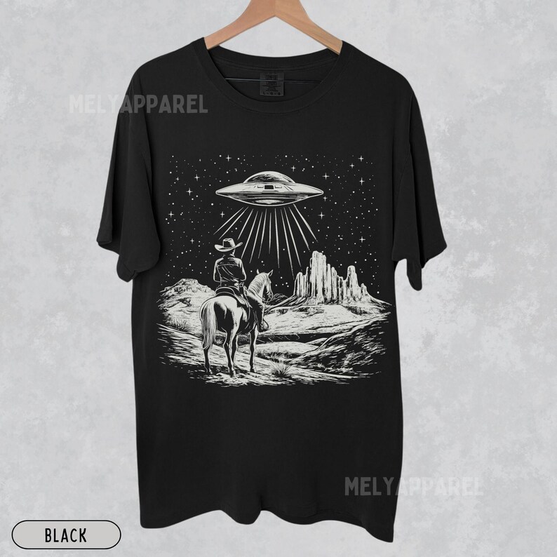 May include: Black t-shirt with a graphic of a cowboy riding a horse under a UFO in a desert landscape.