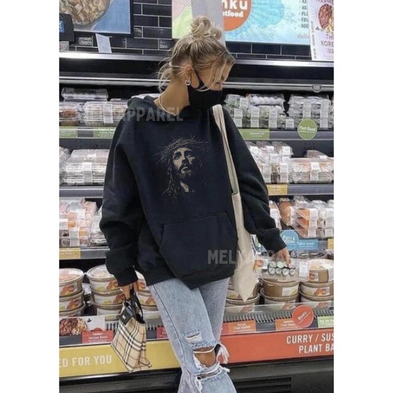 May include: Black hooded sweatshirt with a front graphic of a religious figure wearing a crown of thorns. The sweatshirt has a kangaroo pocket and the word "APPAREL" is printed on the left side. The person is wearing light blue jeans with ripped knees.