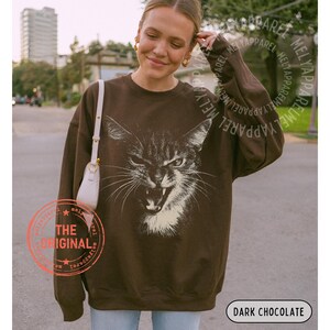 May include: A dark chocolate brown crewneck sweatshirt featuring a large, detailed white graphic print of a cat with an open mouth and bared teeth. The long-sleeved pullover has a relaxed fit and includes a small white shoulder bag strap visible.