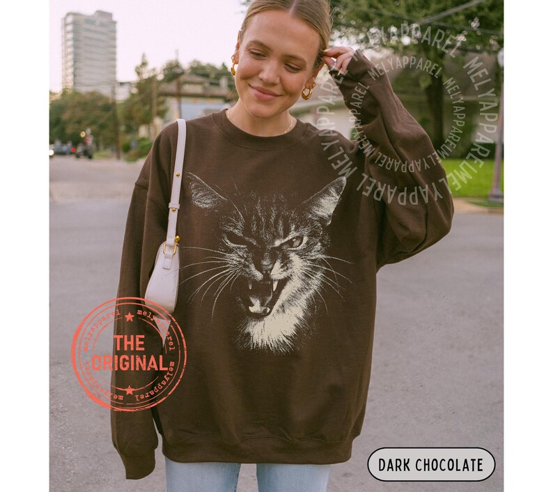 May include: A dark chocolate brown crewneck sweatshirt featuring a large, detailed white graphic print of a cat with an open mouth and bared teeth. The long-sleeved pullover has a relaxed fit and includes a small white shoulder bag strap visible.