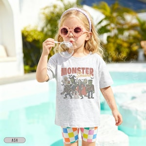 May include: A light gray short-sleeve t-shirt featuring a graphic print of various monsters and the text "MONSTER Season" in brown and black. The shirt is paired with multicolored checkered shorts, a light purple headband, and flower-shaped sunglasses.