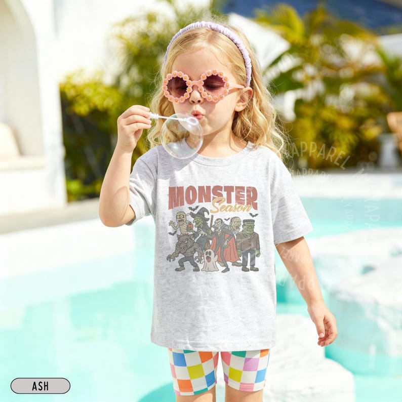 May include: A light gray short-sleeve t-shirt featuring a graphic print of various monsters and the text "MONSTER Season" in brown and black. The shirt is paired with multicolored checkered shorts, a light purple headband, and flower-shaped sunglasses.