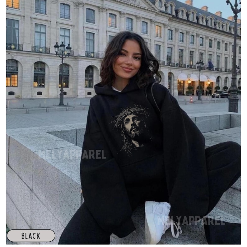 May include: Black hoodie and sweatpants set featuring a graphic of Jesus wearing a crown of thorns. The hoodie has a drawstring hood and a front pocket. The text "MELYAPPAREL" is visible on the clothing. The set is paired with white sneakers.