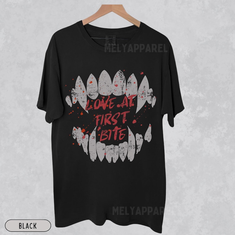K&ouml;nnte beinhalten: Schwarzes T-Shirt mit einer Grafik von Vampirz&auml;hnen und den Worten "LOVE AT FIRST BITE" in Rot. Das Design enth&auml;lt rote Spritzer. Das T-Shirt wird auf einem Holzb&uuml;gel pr&auml;sentiert.