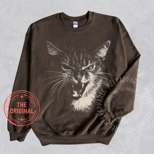 May include: A dark brown crewneck sweatshirt featuring a large, off-white graphic illustration of a hissing cat with bared teeth. A circular red stamp on the lower left reads THE ORIGINAL, surrounded by the repeated text MELYAPPAREL.