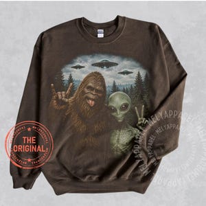 May include: A dark brown crewneck sweatshirt featuring a graphic print of Bigfoot and a green alien standing in a forest with flying saucers in the sky. Bigfoot is making a rock-and-roll hand gesture, and the alien is making a peace sign.