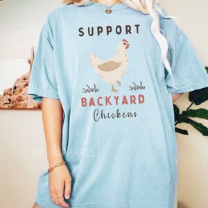 以下が含まれることがあります： 薄い青色のTシャツに、茶色と赤色の文字で「SUPPORT BACKYARD CHICKENS」と書かれています。Tシャツの中央には、漫画の鶏が描かれています。