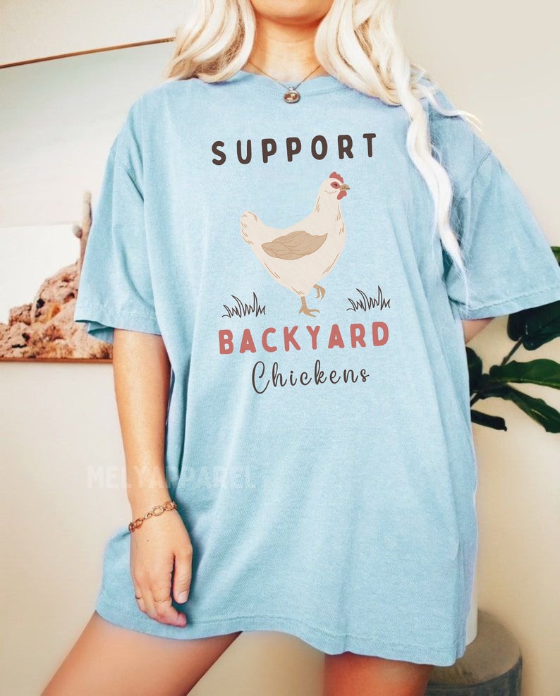 以下が含まれることがあります： 薄い青色のTシャツに、茶色と赤色の文字で「SUPPORT BACKYARD CHICKENS」と書かれています。Tシャツの中央には、漫画の鶏が描かれています。