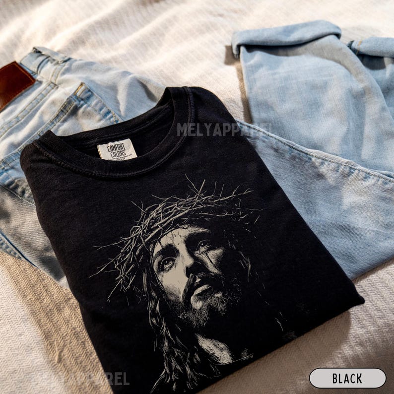 May include: Black t-shirt featuring a detailed grayscale illustration of Jesus wearing a crown of thorns. The shirt is folded, with the text "BLACK" visible. The shirt is placed on a light-colored surface with blue jeans.