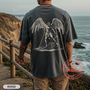 May include: Pepper-colored t-shirt with a large graphic on the back featuring a detailed illustration of an angel with large wings and a sword. The shirt has a relaxed fit and short sleeves. The text "THE ORIGINAL" is printed on the lower right.