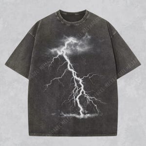May include: Dark gray t-shirt featuring a white lightning bolt graphic. The shirt has a crew neck and short sleeves. The lightning graphic is centered on the front of the shirt, with a cloudy background.