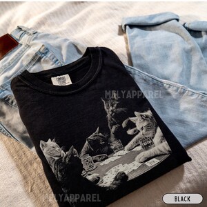 Puede incluir: Camiseta negra con un gr&aacute;fico en escala de grises de gatos jugando al p&oacute;quer. La camiseta tiene cuello redondo y el nombre de la marca "MELYAPPAREL" est&aacute; impreso. La camiseta est&aacute; doblada sobre unos vaqueros azul claro.