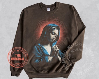 May include: A dark brown crewneck sweatshirt featuring a graphic print of a figure with a head covering and clasped hands, surrounded by a red stippled halo. The text THE ORIGINAL is printed inside a red circular stamp on the lower left side.