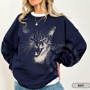 May include: A navy blue crewneck sweatshirt featuring a white graphic illustration of a cat with an open mouth and bared teeth. The design is centered on the chest, and the garment is layered over a white shirt. The text MELYAPPAREL is visible.