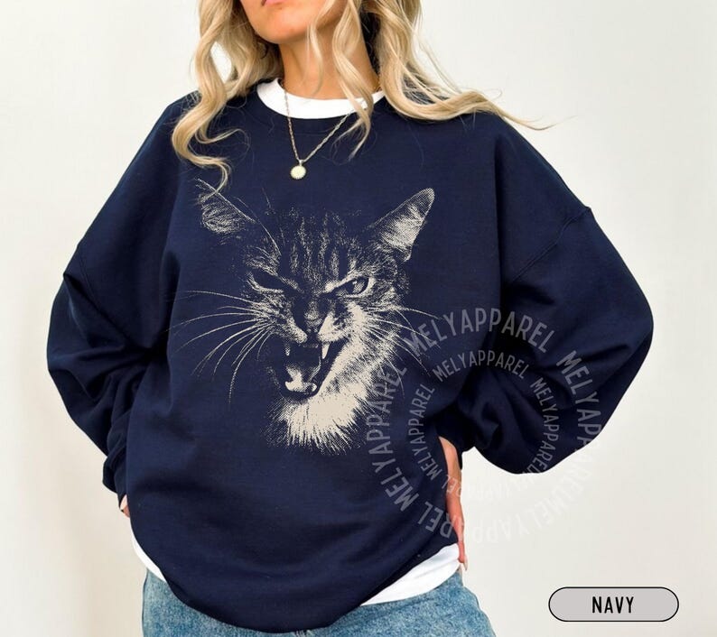 May include: A navy blue crewneck sweatshirt featuring a white graphic illustration of a cat with an open mouth and bared teeth. The design is centered on the chest, and the garment is layered over a white shirt. The text MELYAPPAREL is visible.