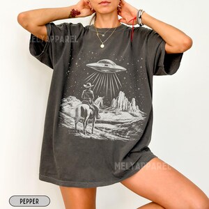 May include: A dark gray oversized t-shirt with a white graphic print of a cowboy riding a horse and a UFO in the sky. The graphic is set against a starry night sky with mountains in the background.