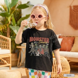 May include: A black short-sleeve t-shirt featuring a graphic print of various monsters and the text "MONSTER Season" in a vintage-style font. The design includes illustrations of a mummy, a witch, a vampire, a werewolf, a ghost, and Frankenstein.