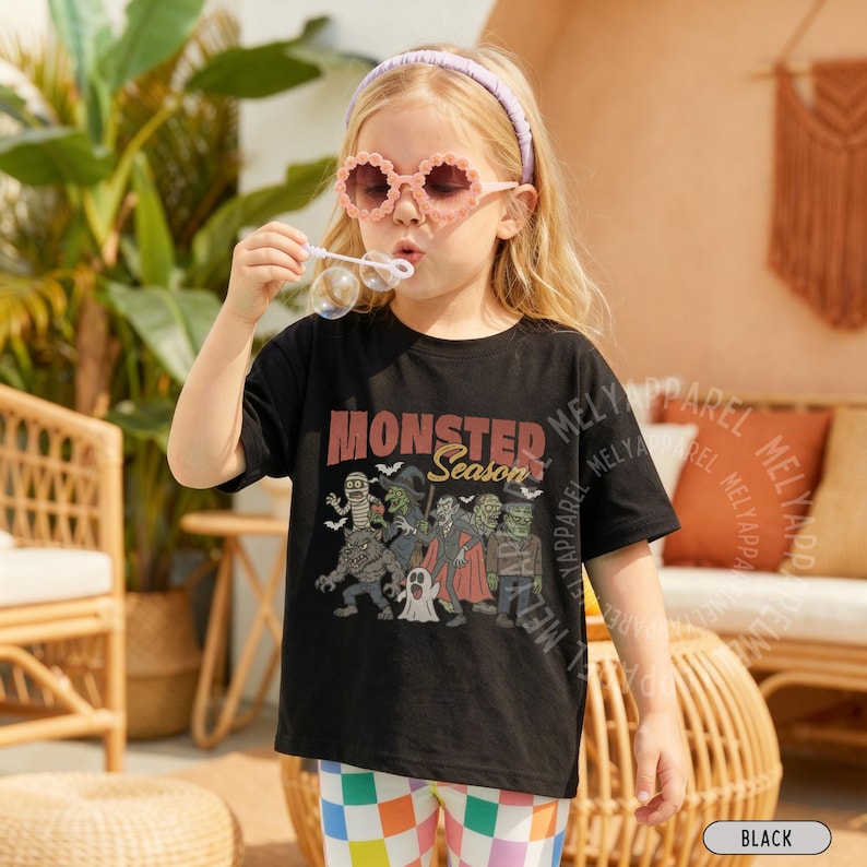 May include: A black short-sleeve t-shirt featuring a graphic print of various monsters and the text "MONSTER Season" in a vintage-style font. The design includes illustrations of a mummy, a witch, a vampire, a werewolf, a ghost, and Frankenstein.