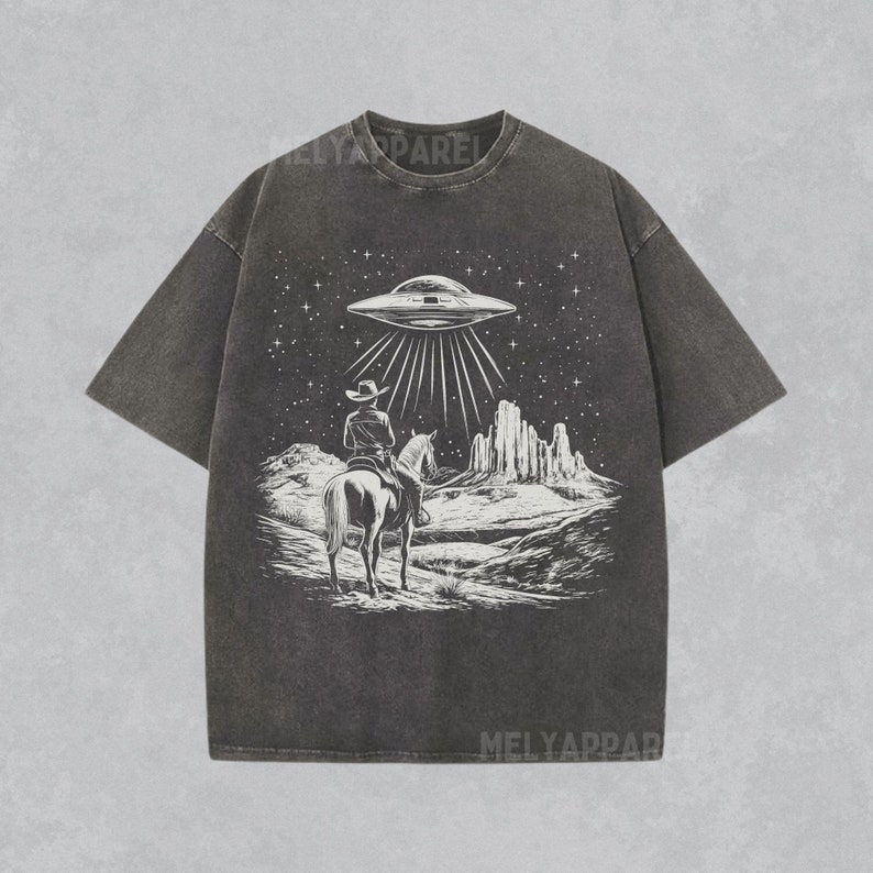 May include: A dark gray t-shirt with a graphic print of a cowboy riding a horse in a desert landscape with a UFO flying overhead. The graphic is in white.