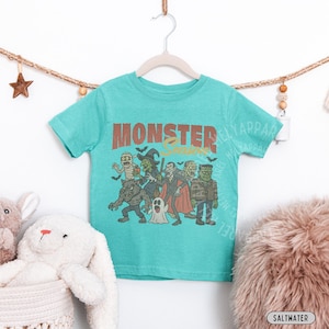 May include: A teal short-sleeve t-shirt featuring a graphic print of classic monsters including a mummy, witch, vampire, werewolf, ghost, and Frankenstein's monster. The text "MONSTER Season" is printed in a bold, distressed brown font above.