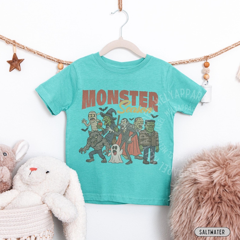 May include: A teal short-sleeve t-shirt featuring a graphic print of classic monsters including a mummy, witch, vampire, werewolf, ghost, and Frankenstein's monster. The text "MONSTER Season" is printed in a bold, distressed brown font above.