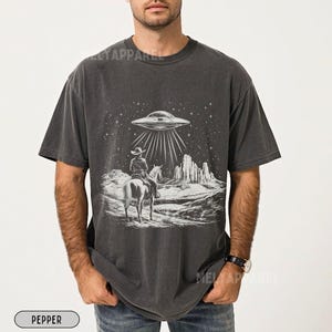 May include: A dark gray t-shirt with a white graphic of a cowboy riding a horse in front of a desert landscape with a UFO hovering above. The graphic also includes stars and a mountain range.