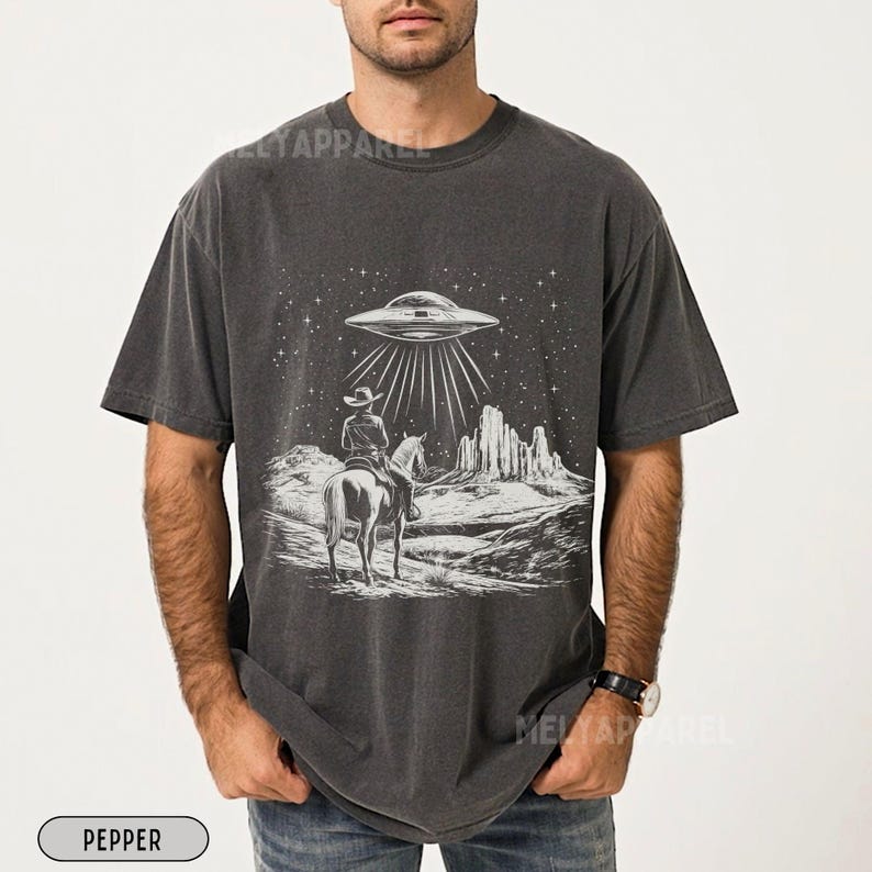 May include: A dark gray t-shirt with a white graphic of a cowboy riding a horse in front of a desert landscape with a UFO hovering above. The graphic also includes stars and a mountain range.