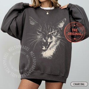 May include: A charcoal gray crewneck sweatshirt featuring a large, white, detailed graphic of a hissing cat face. The design includes a circular orange stamp with the text THE ORIGINAL and a repeating watermark of the brand name MELYAPPAREL.