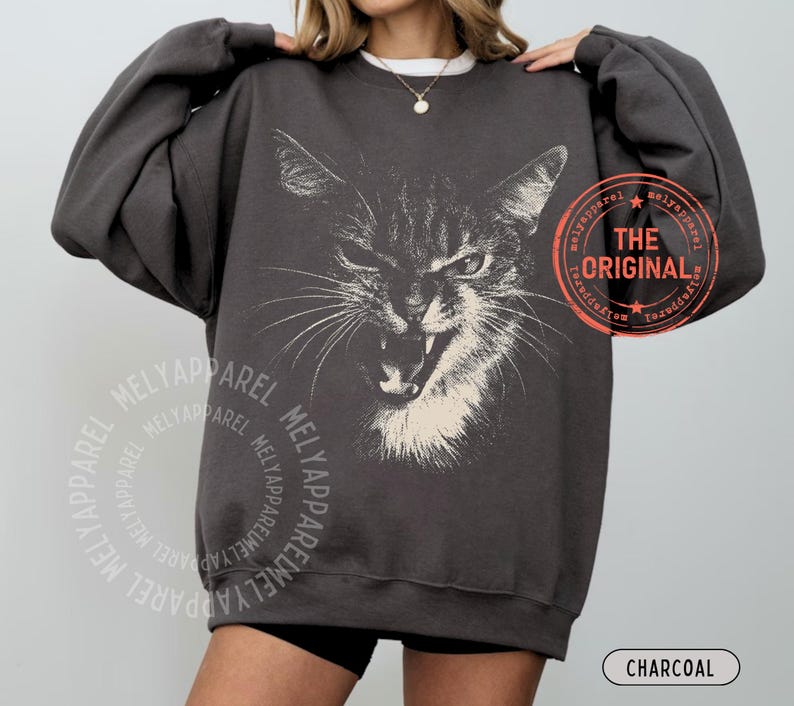 May include: A charcoal gray crewneck sweatshirt featuring a large, white, detailed graphic of a hissing cat face. The design includes a circular orange stamp with the text THE ORIGINAL and a repeating watermark of the brand name MELYAPPAREL.