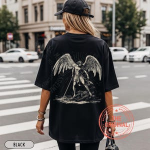 May include: Black oversized t-shirt featuring a detailed graphic of an angel with large wings and a sword. The design is in shades of gray and white. The word "BLACK" is printed at the bottom.