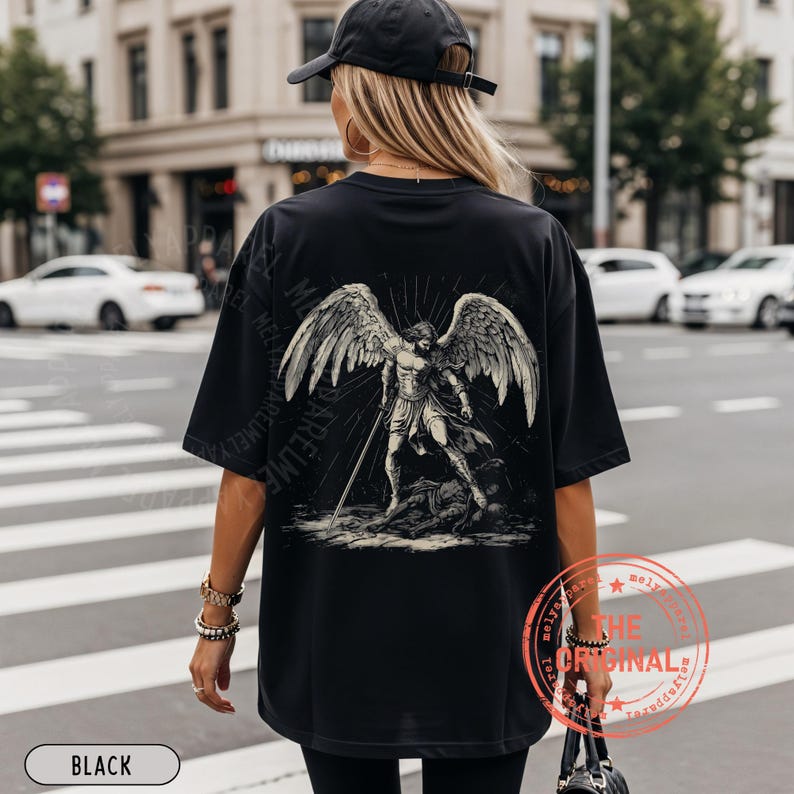May include: Black oversized t-shirt featuring a detailed graphic of an angel with large wings and a sword. The design is in shades of gray and white. The word "BLACK" is printed at the bottom.