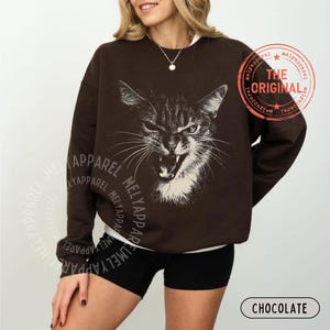 May include: A dark brown long-sleeve sweatshirt featuring a white graphic illustration of a cat with an open mouth and bared teeth. The text "THE ORIGINAL" is printed in a circular red stamp design on the upper right side of the garment.